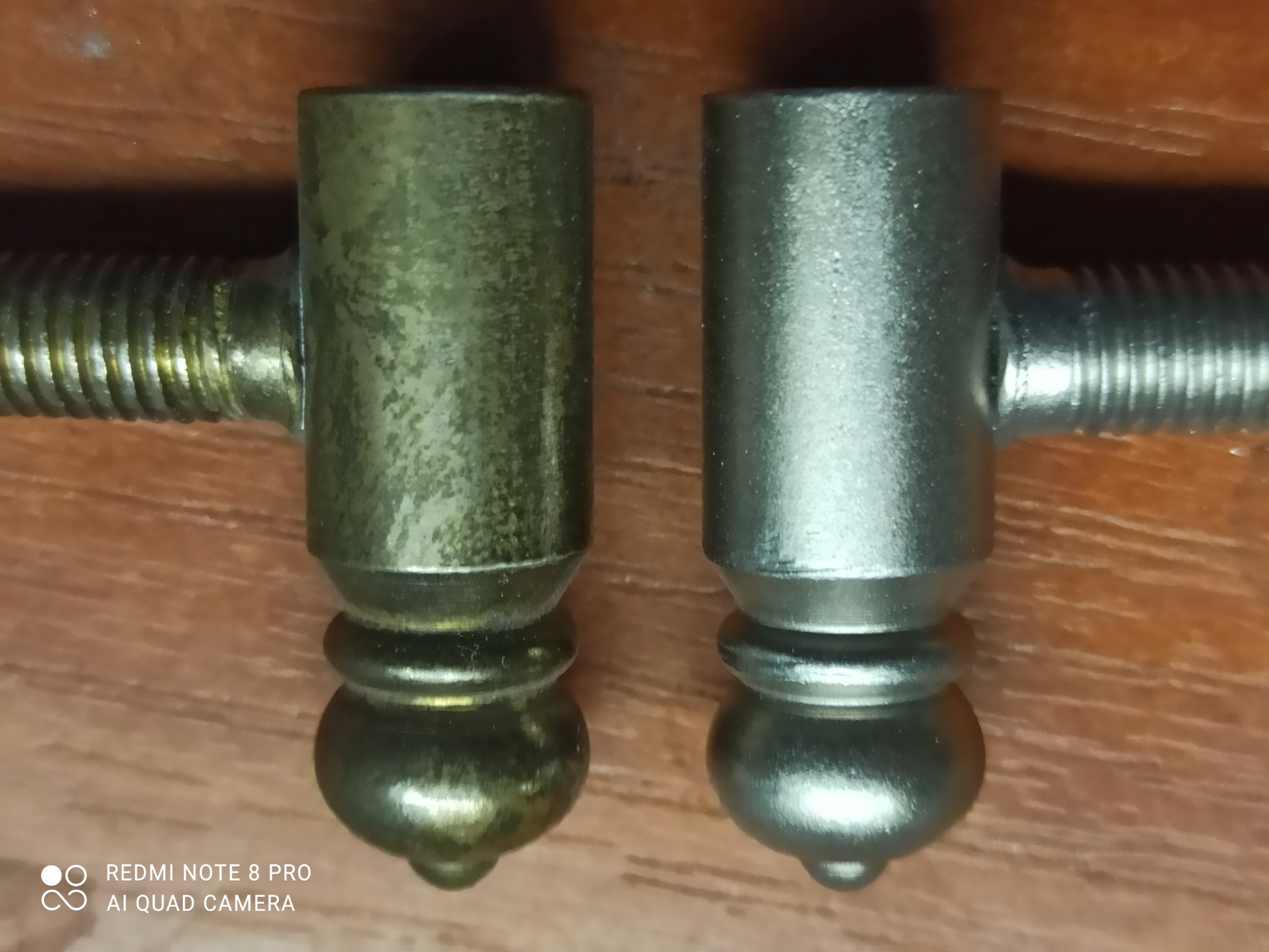 Dwa metalowe elementy z gwintem, jeden w odcieniu starego złota, drugi srebrny, leżące na drewnianej powierzchni. Element po lewej stronie wygląda na starszy lub zniszczony.