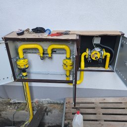 INSTALACJE Patryk Sędziński - Żółta instalacja gazowa w brązowej szafce zewnętrznej z otwartymi drzwiczkami, widoczne narzędzia na górze szafki, paleta drewniana i kanister pod spodem, świeżo wykopana ziemia wokół.