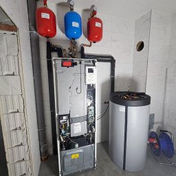INSTALACJE Patryk Sędziński - Wnętrze kotłowni z otwartym panelem sterowania kotła grzewczego, trzema zbiornikami wyrównawczymi (dwa czerwone, jeden niebieski) pod sufitem, zasobnikiem ciepłej wody z narzędziami na górze...
