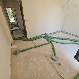 INSTALACJE Patryk Sędziński - Wnętrze w trakcie montażu rekuperacji: zielone, karbowane rury wentylacyjne rozłożone na surowej podłodze, widoczne elementy montażowe i fragment okna.