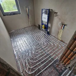 Rozłożona instalacja ogrzewania podłogowego na folii izolacyjnej z kratką, widoczny stelaż podtynkowy WC Geberit, ściany w stanie surowym, instalacje wodne zabezpieczone pianką montażową.