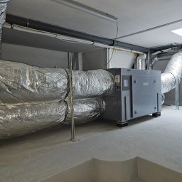 INSTALACJE Patryk Sędziński - Szare urządzenie wentylacyjne Pro-Vent Mistral Pro 1400 z podłączonymi izolowanymi przewodami wentylacyjnymi w pomieszczeniu technicznym z betonową podłogą.