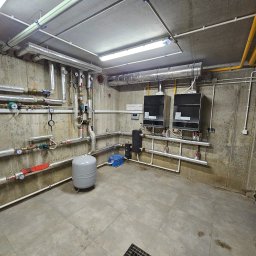 INSTALACJE Patryk Sędziński - Węzeł cieplny w piwnicy: dwa piece grzewcze, zbiornik wyrównawczy, liczne rury miedziane i stalowe z zaworami i manometrami, betonowe ściany i podłoga z płytek.