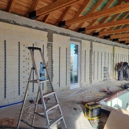 INSTALACJE Patryk Sędziński - Montaż ogrzewania podłogowego wodnego w surowym budynku z drewnianą konstrukcją dachu, widoczna drabina z butelką wody i narzędzia budowlane.