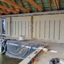 INSTALACJE Patryk Sędziński - Wnętrze w trakcie budowy z widocznymi ścianami z bloczków, na których zamontowano listwy instalacyjne. Pracownik na drabinie instaluje przewody. Część pomieszczenia przykryta folią ochronną.