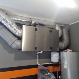 INSTALACJE Patryk Sędziński - Centrala wentylacyjna reQ-net z podłączonymi przewodami wentylacyjnymi, widoczny zbiornik wyrównawczy i bojler, całość na tle szarej ściany z pomarańczowym pasem.