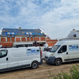 INSTALACJE Patryk Sędziński - Budowa domu w trakcie zadaszenia z widocznymi oknami dachowymi, dwa białe samochody dostawcze firmy AUX Klimatyzatory na pierwszym planie.