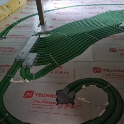 INSTALACJE Patryk Sędziński - Zielone, karbowane rury wentylacyjne rozłożone na izolacji XPS Carbon w poddaszu, widoczne drewniane elementy konstrukcyjne dachu.