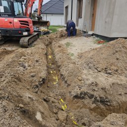 INSTALACJE Patryk Sędziński - Wykopany rów z żółtą taśmą ostrzegawczą, w tle koparka Kubota U55-4 i pracownicy przy budynku w trakcie prac ziemnych.