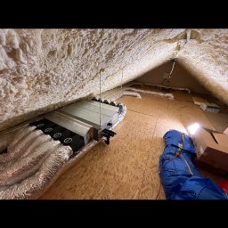 INSTALACJE Patryk Sędziński - Centrala wentylacyjna z podłączonymi przewodami w przestrzeni poddasza, ściany i dach pokryte pianą izolacyjną, widoczne narzędzia i materiały budowlane.