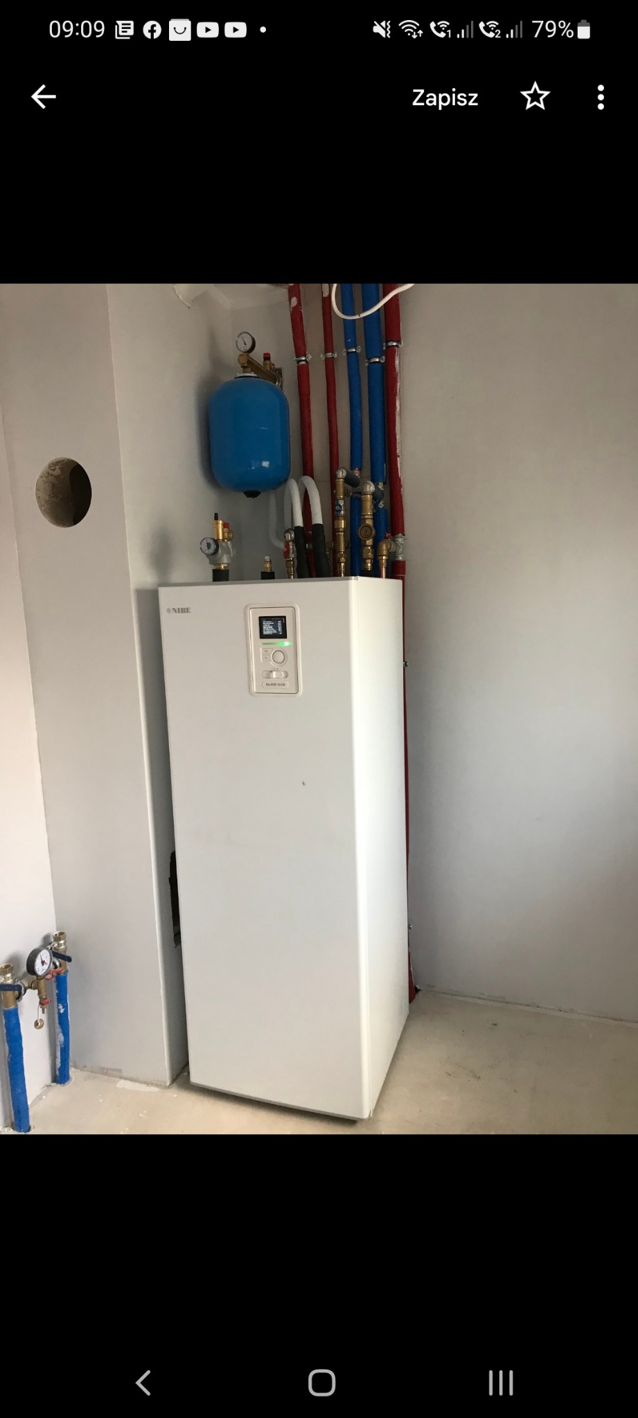 Biała jednostka wewnętrzna urządzenia grzewczego NIBE zamontowana na ścianie, z podłączonymi rurami miedzianymi i niebieskimi, oraz zbiornikiem wyrównawczym, widoczne elementy instalacji...