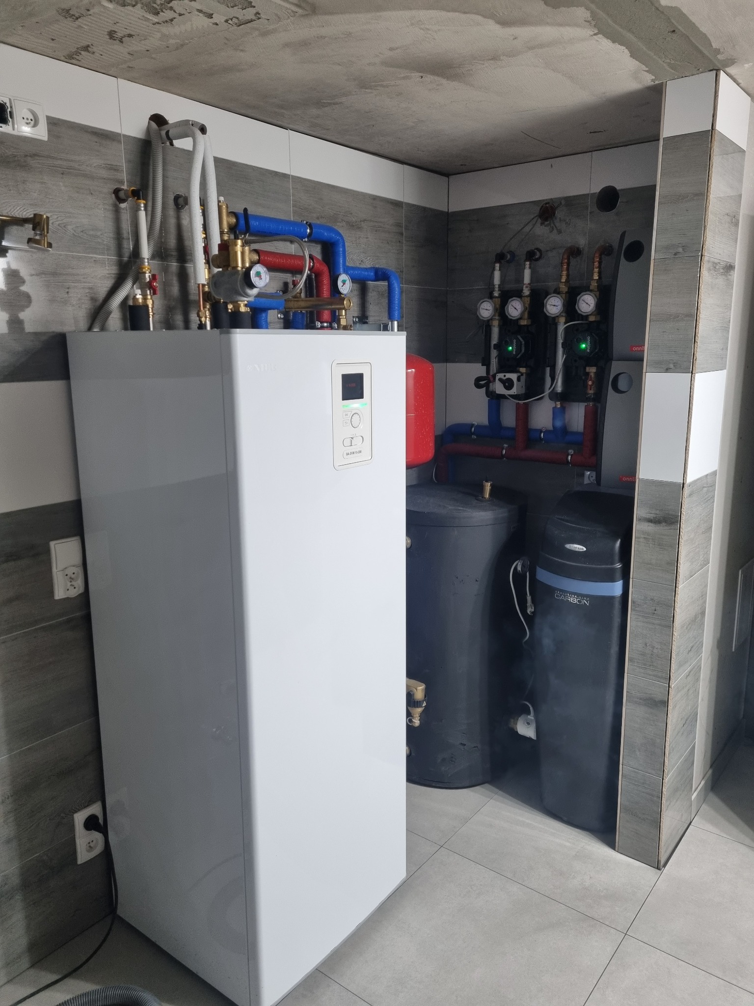 Biała jednostka centralna z rurami w kolorze niebieskim i czerwonym, podłączona do szarego zasobnika i filtra wody w pomieszczeniu wykończonym szarymi płytkami imitującymi drewno, z widoczną surową...