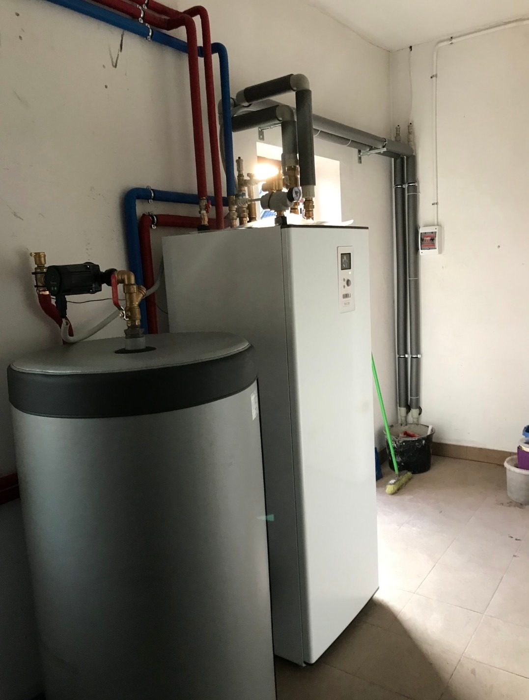Biała pompa ciepła z szarym zasobnikiem ciepłej wody, widoczne rury instalacyjne w kolorze czerwonym i niebieskim, oraz szare rury wentylacyjne pod sufitem w pomieszczeniu technicznym.