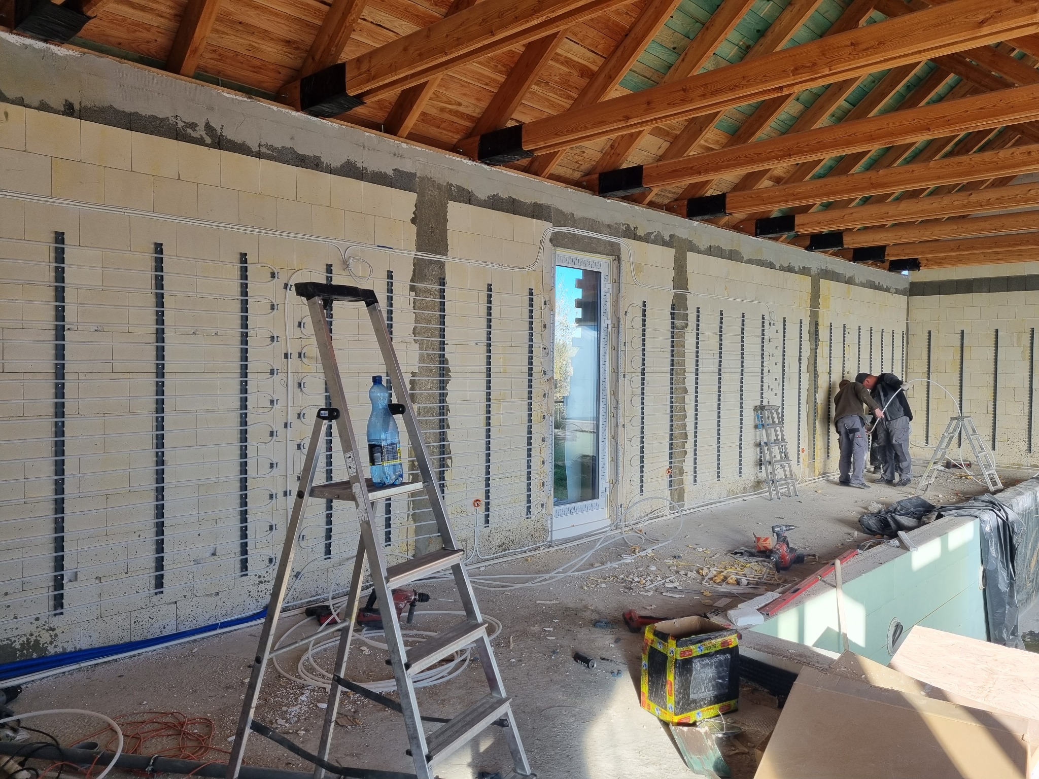 Montaż ogrzewania podłogowego wodnego w surowym budynku z drewnianą konstrukcją dachu, widoczna drabina z butelką wody i narzędzia budowlane.