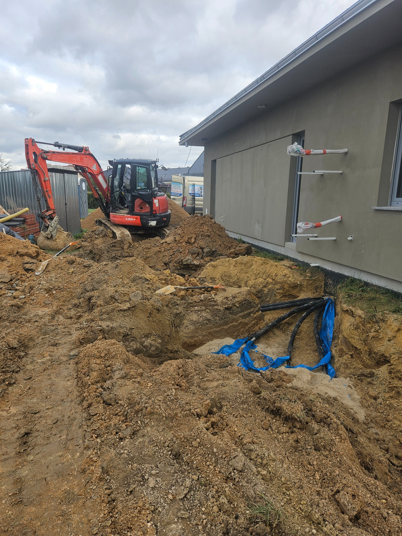 Wykonanie wykopu pod przyłącze wodno-kanalizacyjne obok budynku, widoczny pomarańczowy minikoparka Kubota U55-4, wykop zabezpieczony niebieską folią, wystające rury.