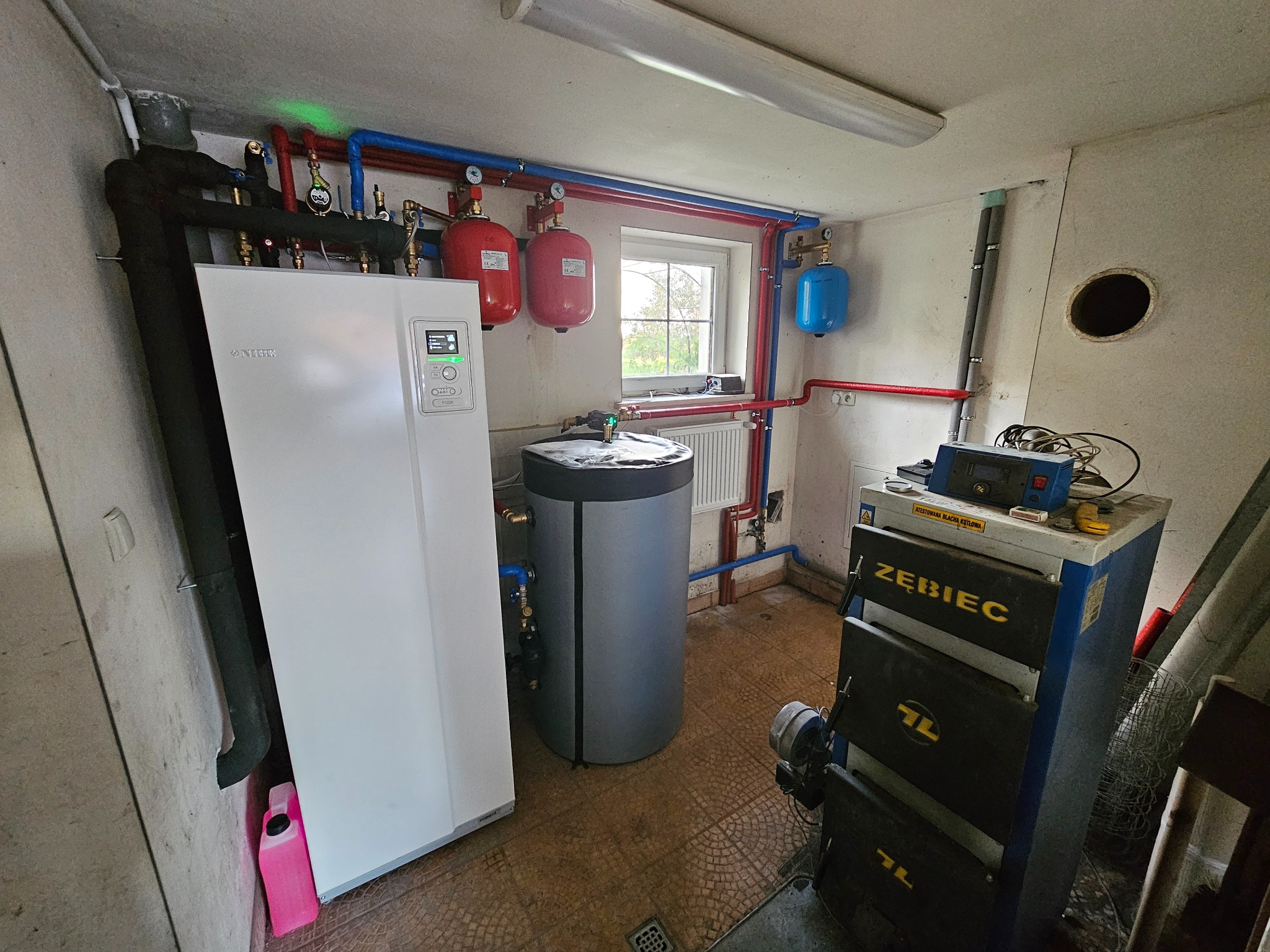 Kotłownia z białą pompą ciepła Nibe, dwoma czerwonymi i jednym niebieskim zbiornikiem ciśnieniowym, szarym zasobnikiem CWU oraz starym piecem węglowym Żębiec. Instalacja z rur miedzianych...