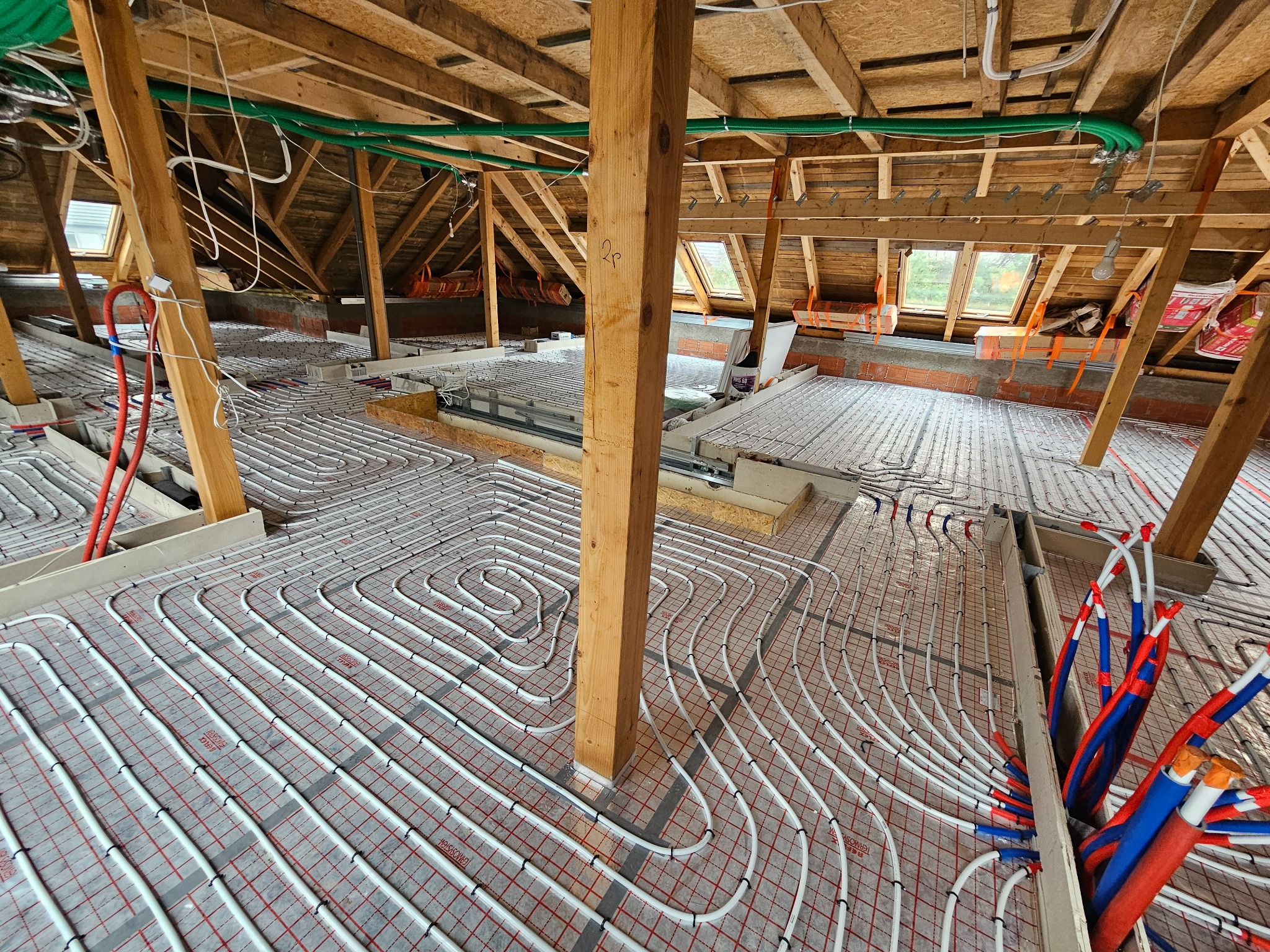 Instalacja ogrzewania podłogowego w trakcie montażu na poddaszu, rury układane spiralnie na izolacji z siatką, widoczne drewniane belki konstrukcyjne i okna dachowe.