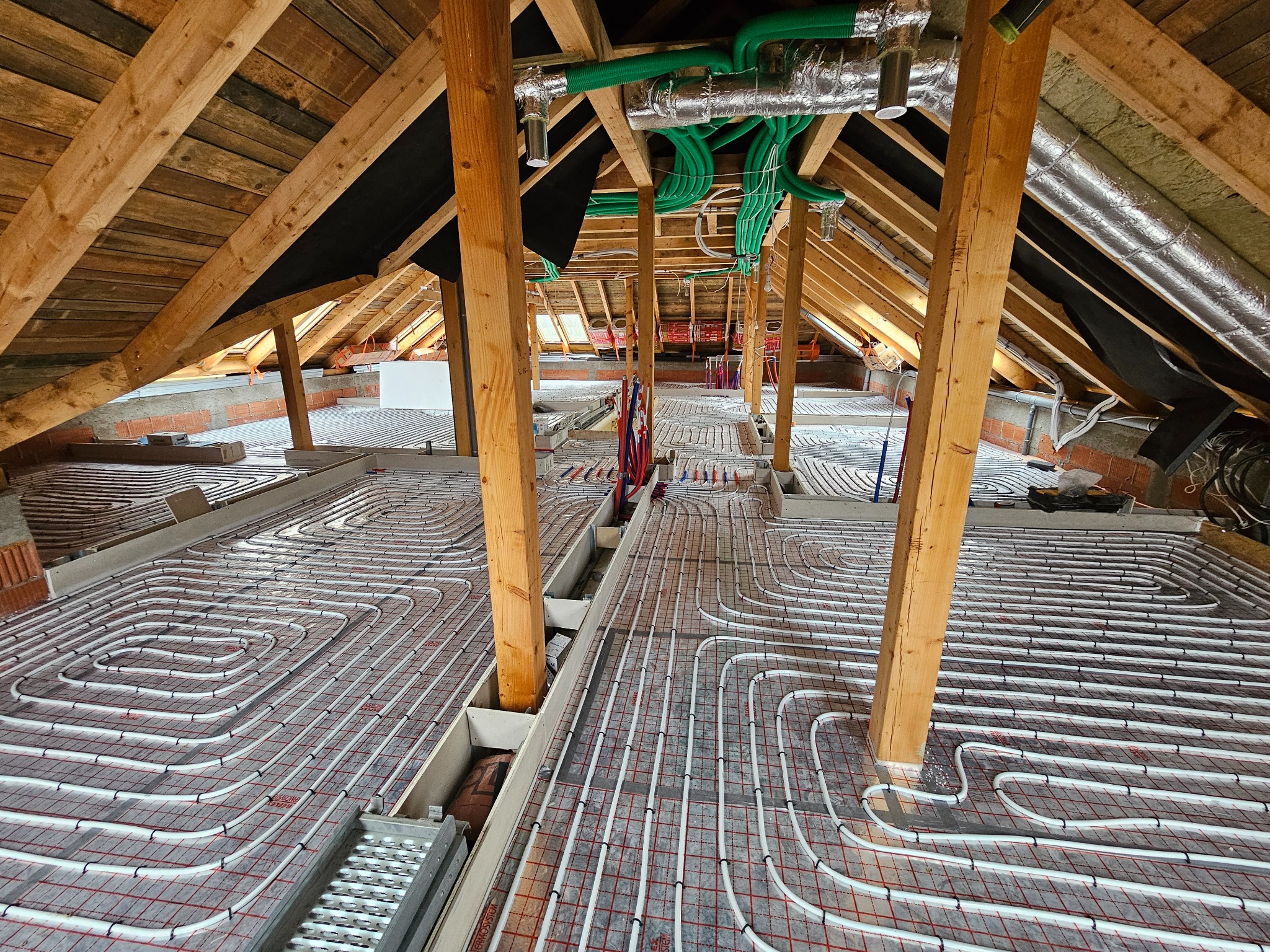 Instalacja ogrzewania podłogowego w trakcie montażu na poddaszu, widoczne rury grzewcze ułożone na izolacji z czerwoną siatką, drewniana konstrukcja dachu i zielone rury wentylacyjne pod sufitem.