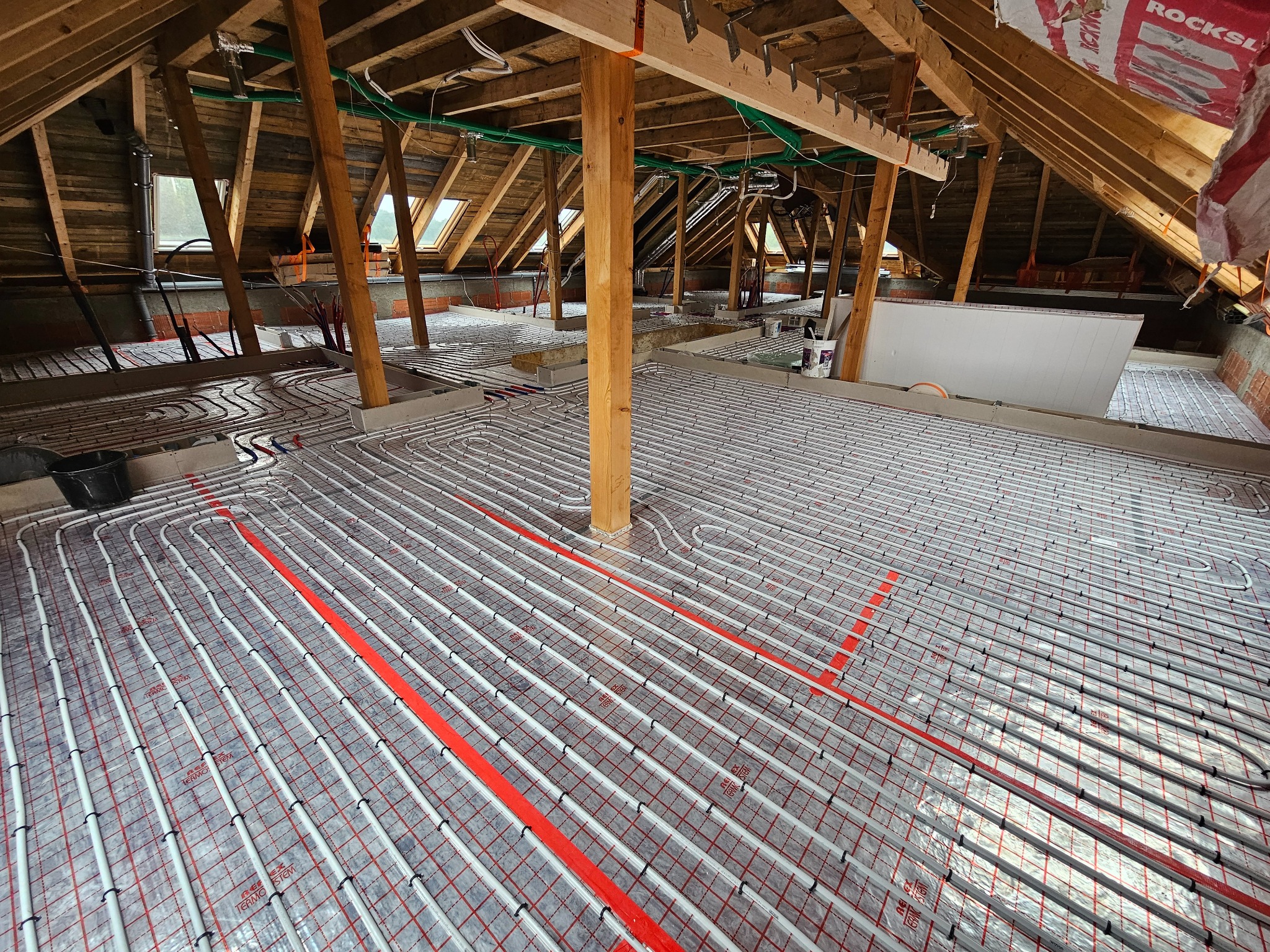 Podłogówka w trakcie montażu na poddaszu z widocznymi belkami stropowymi i oknami dachowymi, rurki grzewcze ułożone w regularnych odstępach na folii z czerwoną siatką.