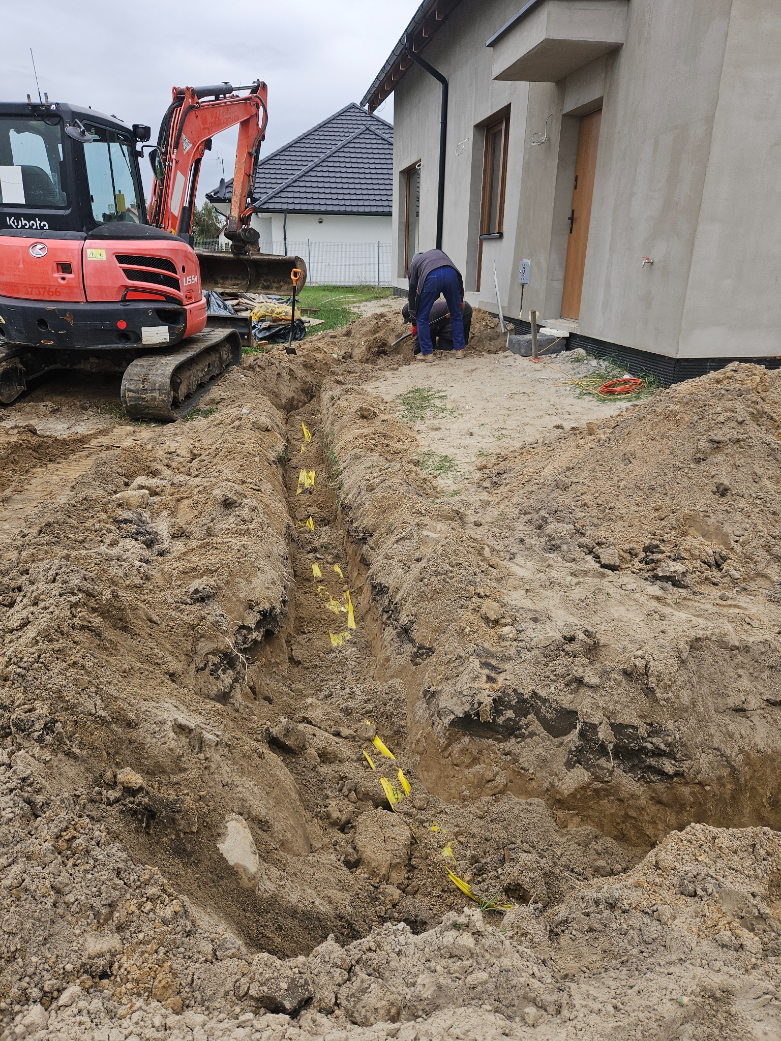 Wykopany rów z żółtą taśmą ostrzegawczą, w tle koparka Kubota U55-4 i pracownicy przy budynku w trakcie prac ziemnych.