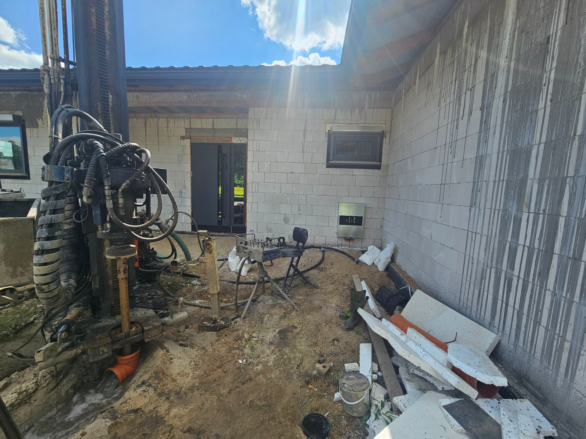 Wiertnica pionowa w trakcie wykonywania odwiertu pod pompę ciepła przy ścianie budynku z betonu komórkowego, widoczne rury i osprzęt wiertniczy oraz elementy instalacji wewnątrz budynku.