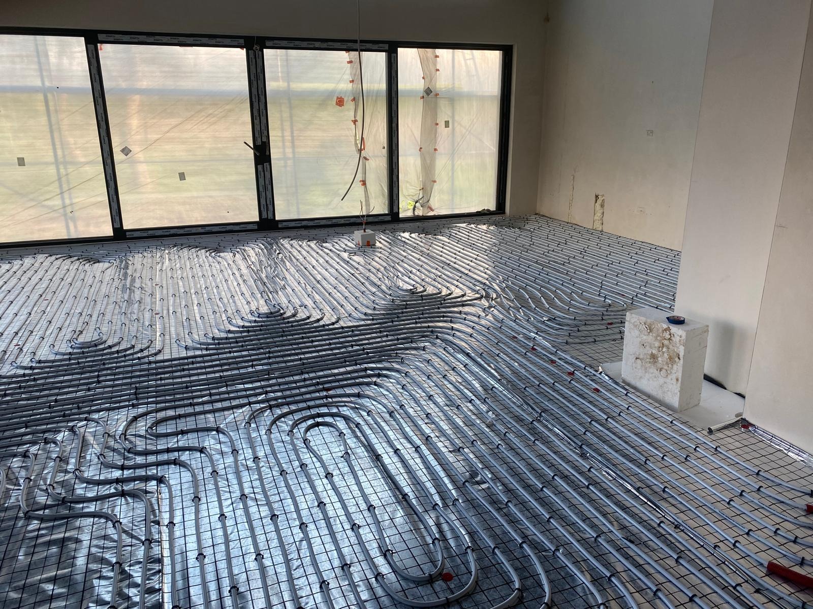 Szare rury ogrzewania podłogowego ułożone na srebrnej folii refleksyjnej w surowym wnętrzu z dużym oknem i betonowym słupem, widoczne elementy instalacji.