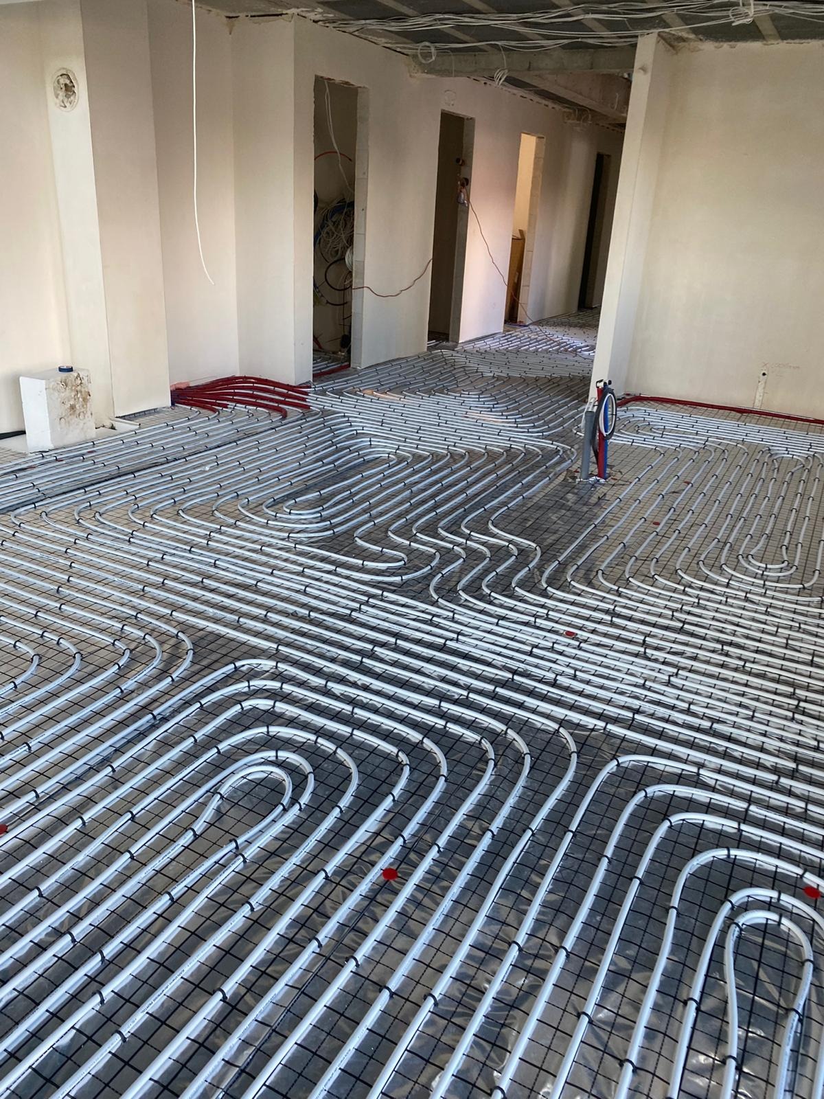 Rozłożona instalacja ogrzewania podłogowego z białych rurek na czarnej siatce w surowym wnętrzu budynku w trakcie budowy, widoczne czerwone elementy i niezabezpieczone kable na suficie.