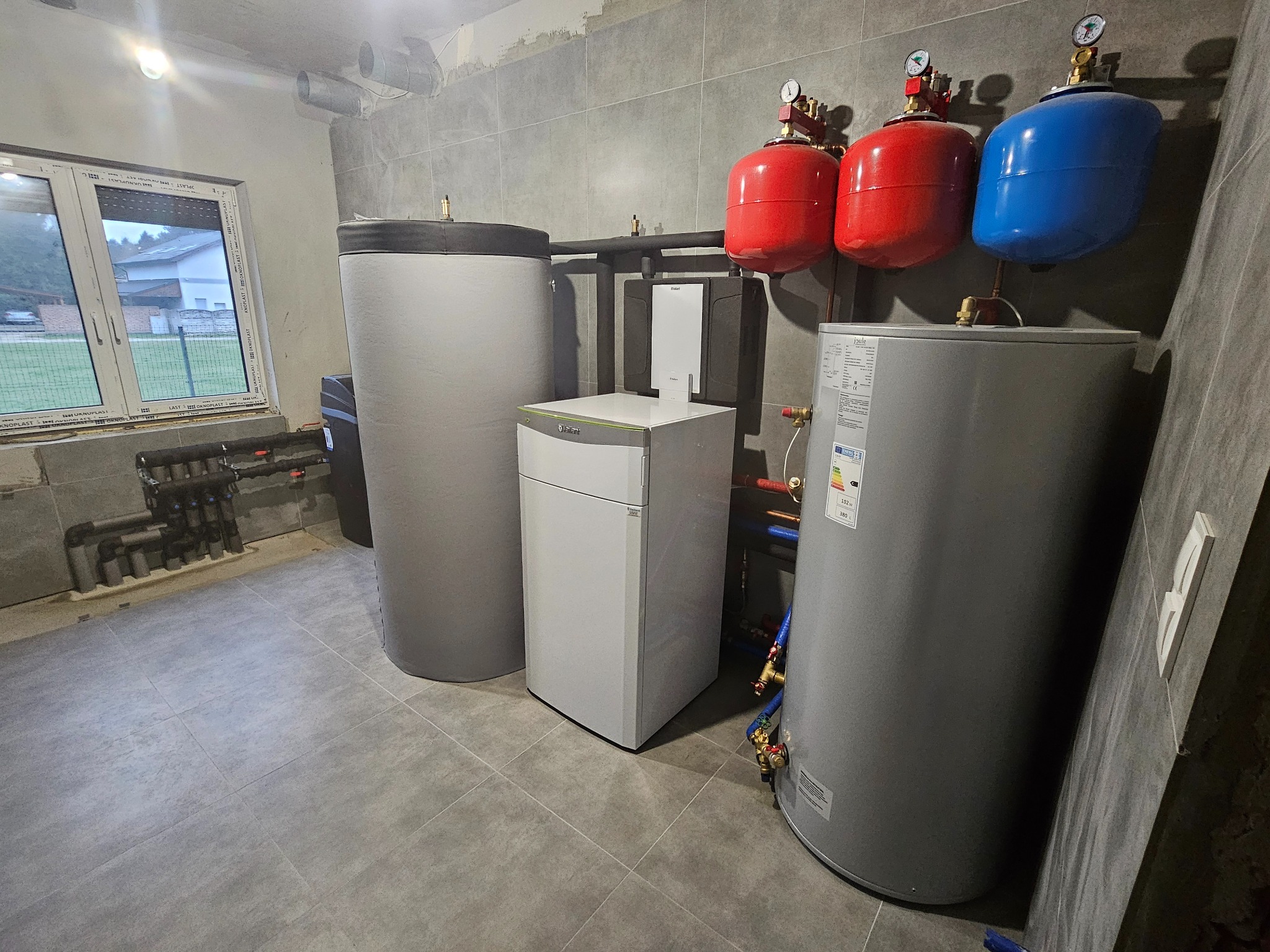 Wnętrze kotłowni z zainstalowanym kotłem grzewczym, dwoma zbiornikami akumulacyjnymi, naczyniami wzbiorczymi w kolorach czerwonym i niebieskim oraz rozdzielaczem hydraulicznym przy oknie.
