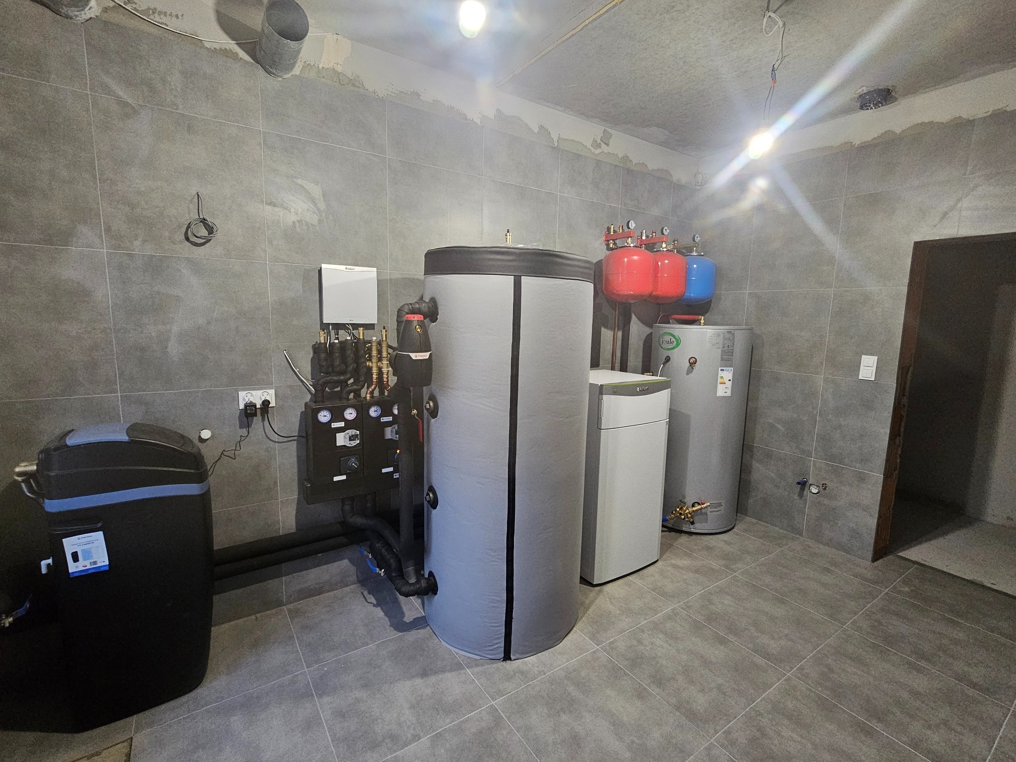 Kompletna instalacja grzewcza w kotłowni z szarymi płytkami podłogowymi i ściennymi, widoczne zasobniki ciepłej wody, zbiornik buforowy w izolacji, grupa pompowa oraz naczynia przeponowe w kolorach...