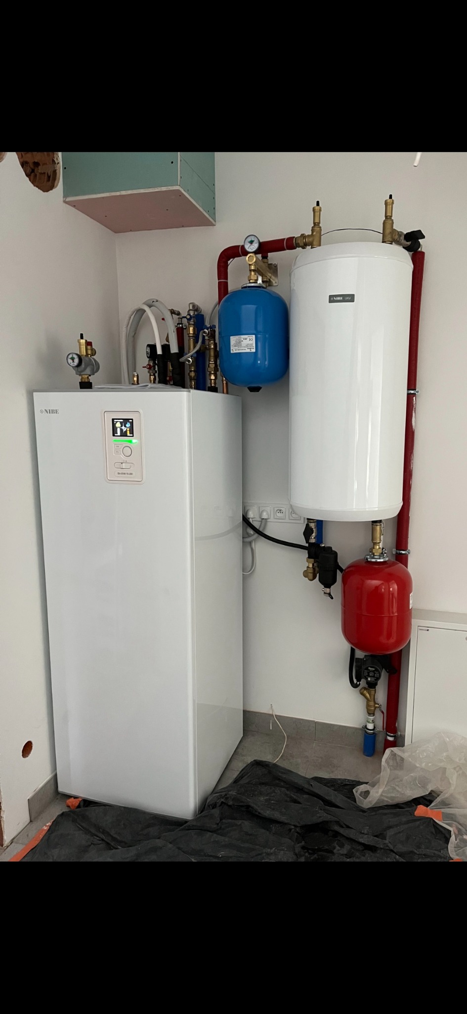 Biała pompa ciepła Nibe z podłączonymi zbiornikami wyrównawczymi (czerwony i niebieski) oraz zasobnikiem ciepłej wody, zainstalowana w pomieszczeniu w Namysłowie, na podłodze rozłożona czarna folia...