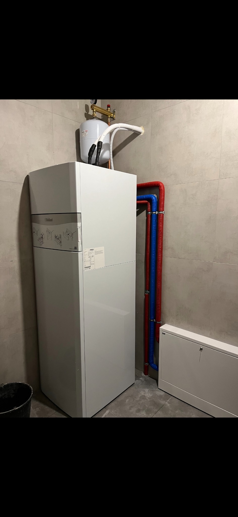 Biała jednostka wewnętrzna z zbiornikiem wyrównawczym, rurami w kolorze czerwonym i niebieskim, zamontowana na tle płytek imitujących beton.