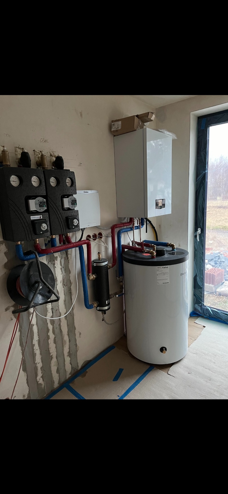 Biały zasobnik ciepłej wody użytkowej z podłączonymi rurami miedzianymi i niebieskimi, zamontowany w surowym pomieszczeniu z widocznymi śladami po tynku i zabezpieczeniem podłogi taśmą malarską.