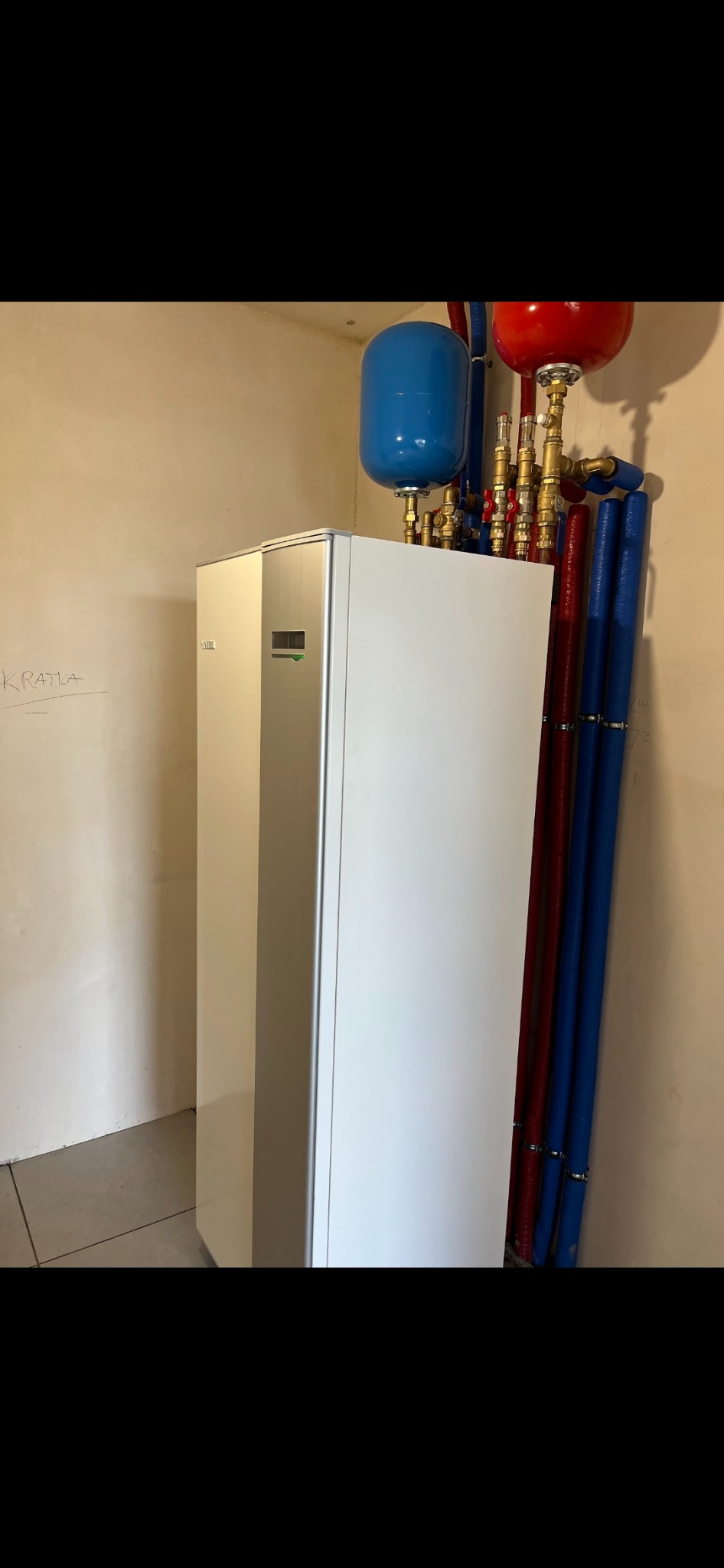 Zainstalowana pompa ciepła Nibe w kotłowni, widoczne rury w izolacji niebieskiej i czerwonej, zbiorniki wyrównawcze, ściana z napisem 'Kratka'.