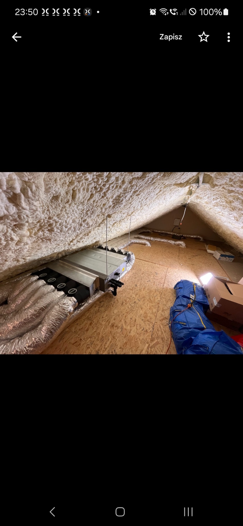 Centrala wentylacyjna z podłączonymi przewodami w przestrzeni poddasza, ściany i dach pokryte pianą izolacyjną, widoczne narzędzia i materiały budowlane.