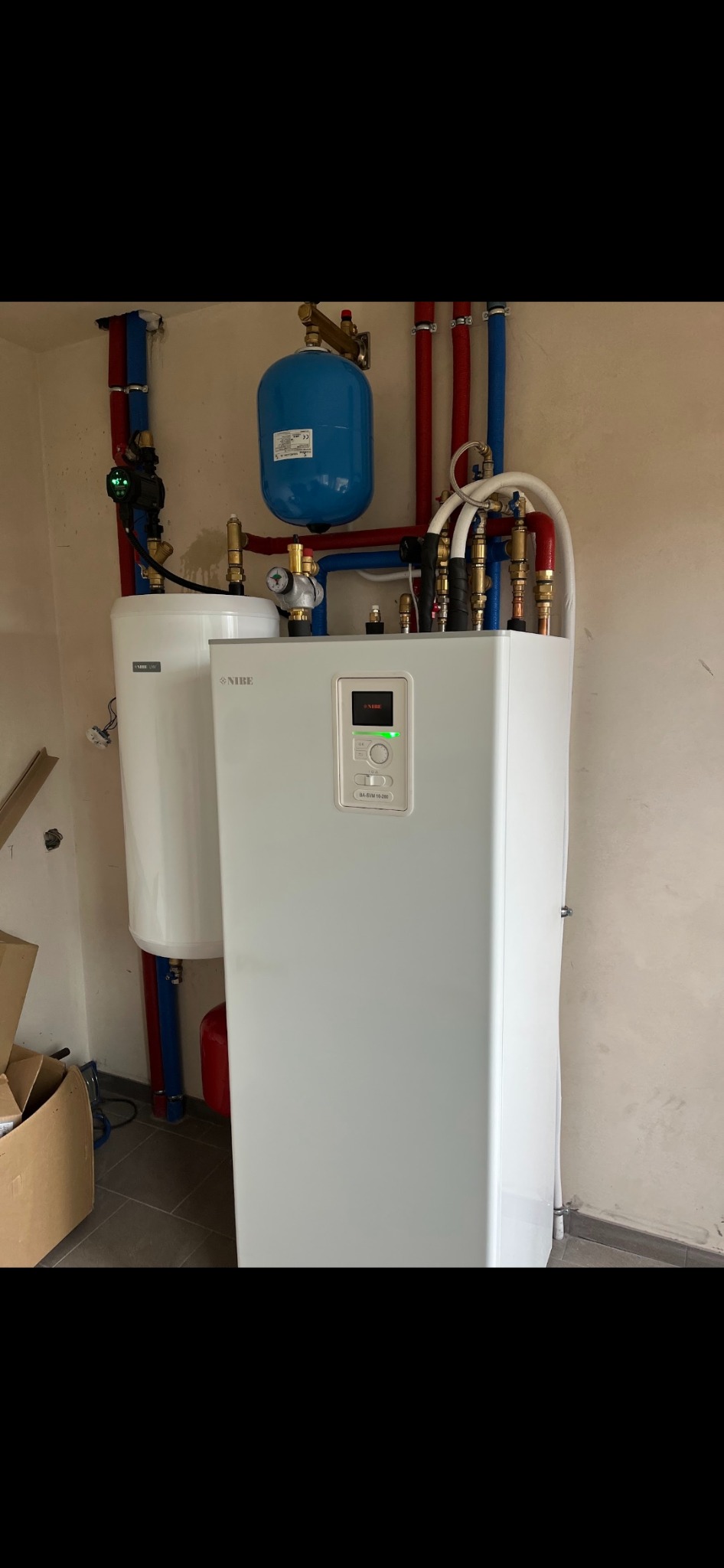 Biała pompa ciepła Nibe z podłączonymi rurami w kolorach czerwonym i niebieskim, zbiornik wyrównawczy koloru niebieskiego, widoczna instalacja hydrauliczna.