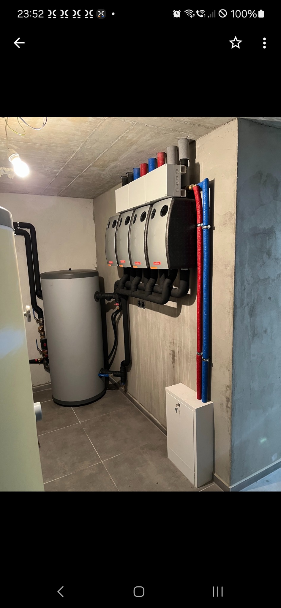 Rozdzielacz podłogowy z czterema obiegami grzewczymi, zbiornik buforowy, rury w izolacji termicznej, instalacja w surowym pomieszczeniu z betonowym sufitem i szarą podłogą z płytek.