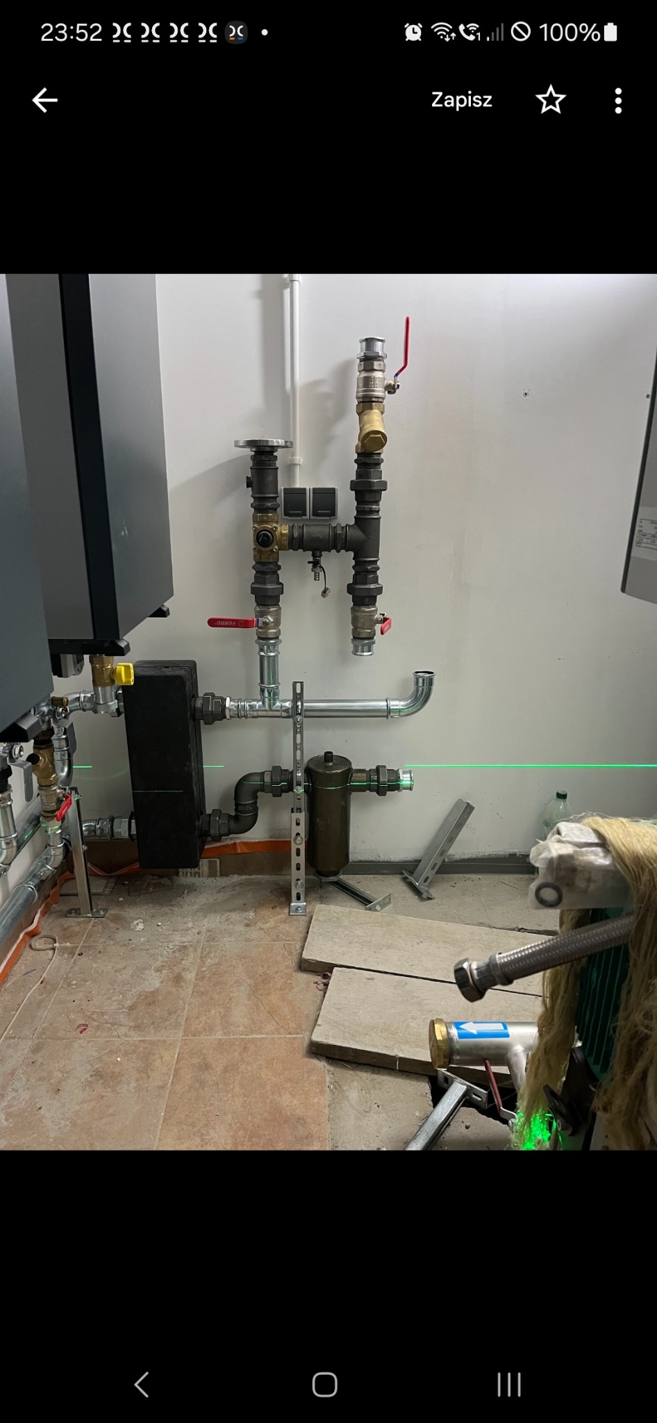 Instalacja grzewcza w kotłowni z czarnym kotłem, rurami, zaworami i filtrem, poziomowana zielonym laserem na brązowych płytkach podłogowych.