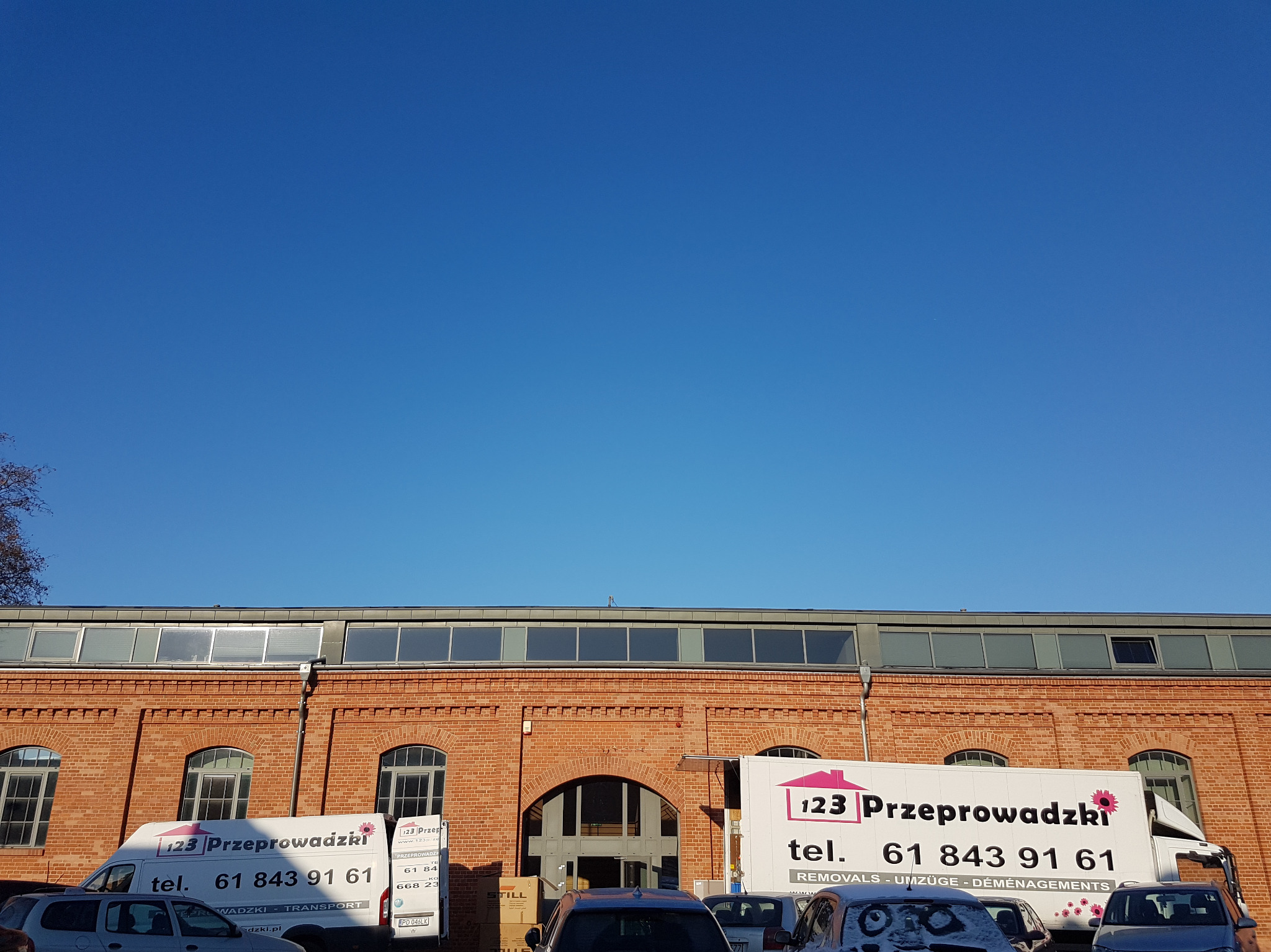 Dwa białe busy z logo firmy przeprowadzkowej zaparkowane przed ceglanym budynkiem z wysokimi oknami i błękitnym niebem w tle.