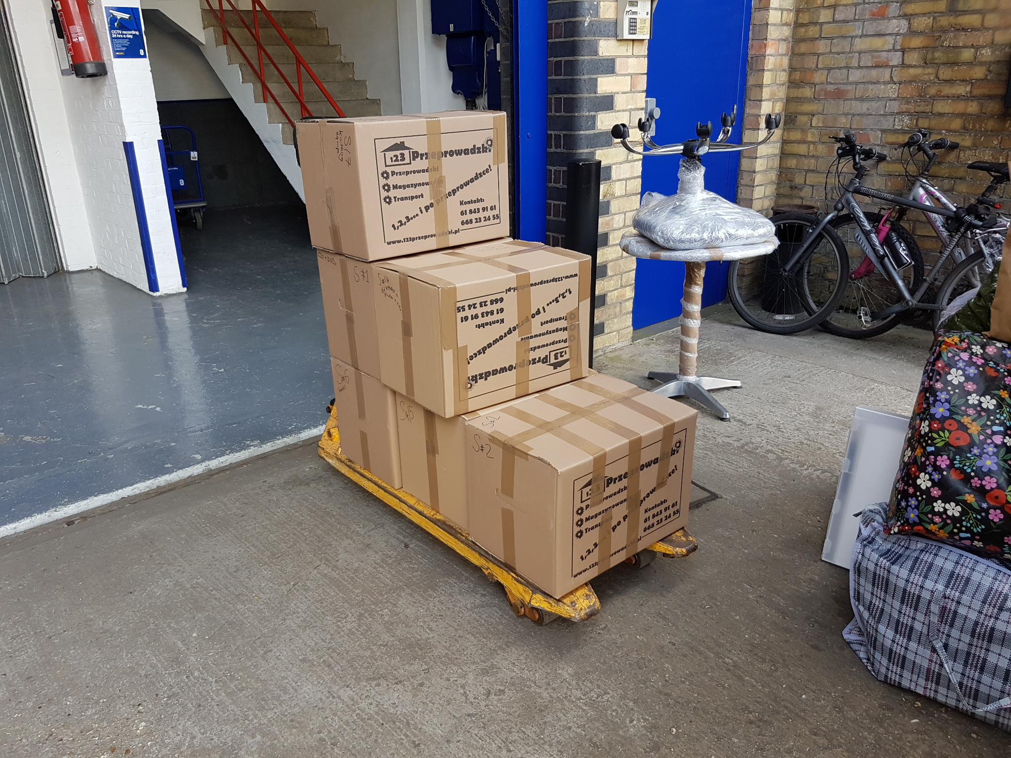 Stos trzech kartonowych pudeł z nadrukowanym logo firmy przeprowadzkowej, umieszczonych na żółtym wózku paletowym w magazynie lub garażu, obok zaparkowanych rowerów i krzesła zabezpieczonego folią.