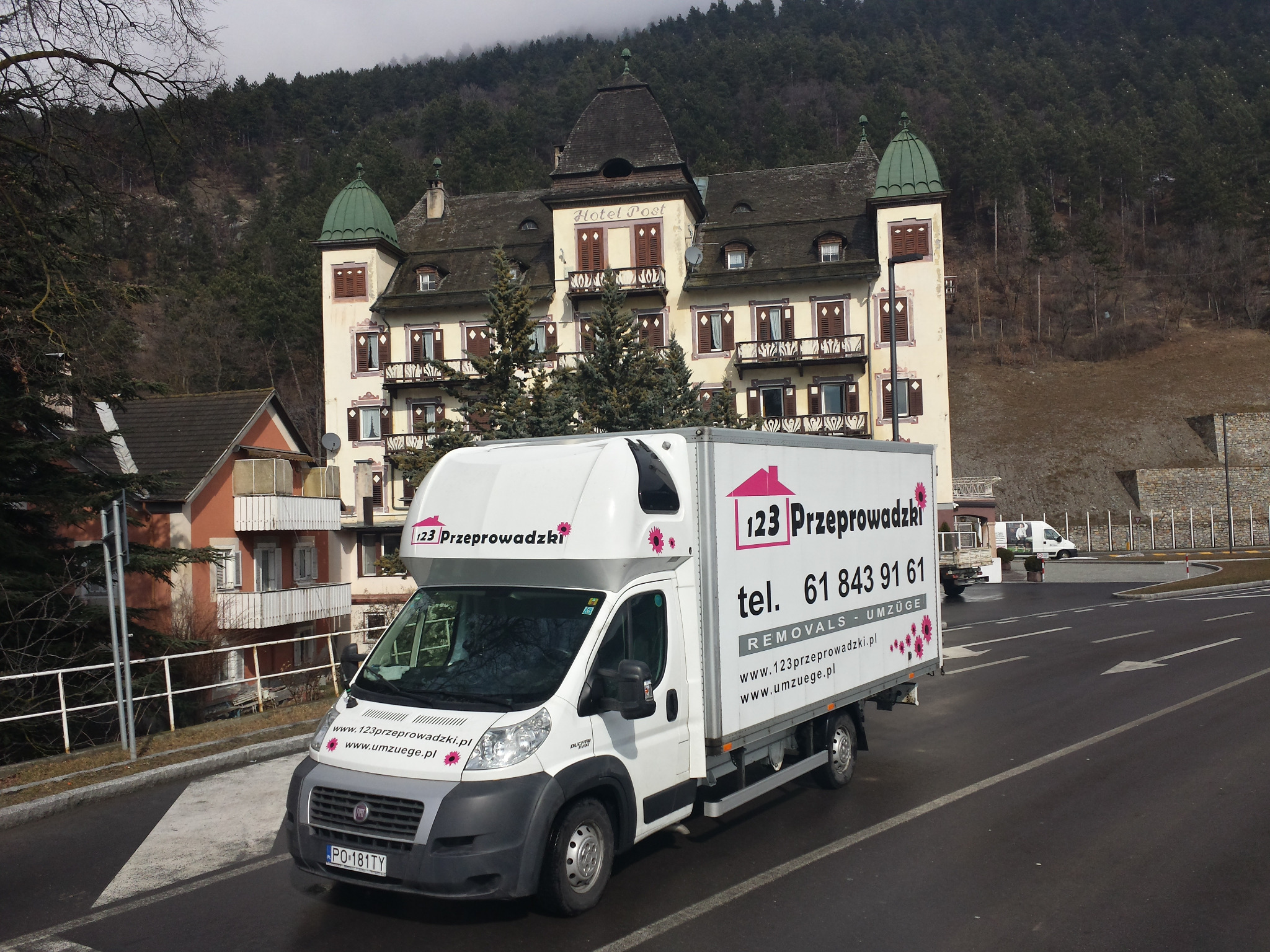 Biała furgonetka z logo firmy przeprowadzkowej na tle zabytkowego hotelu 'Hotel Post' i zalesionych wzgórz.