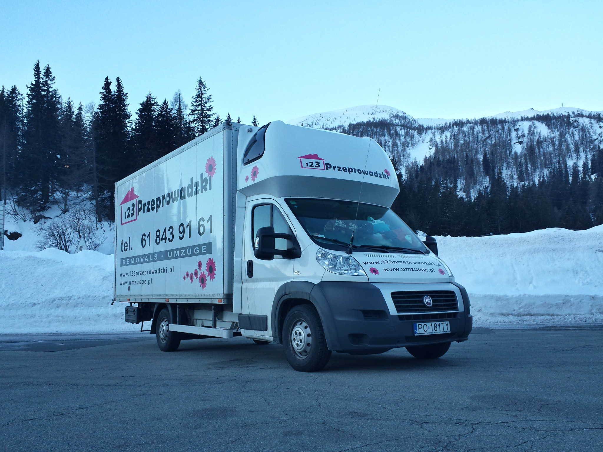 Samochód dostawczy firmy z Poznania, oklejony reklamą usług transportowych i przeprowadzkowych, zaparkowany na tle górskiego krajobrazu pokrytego śniegiem.