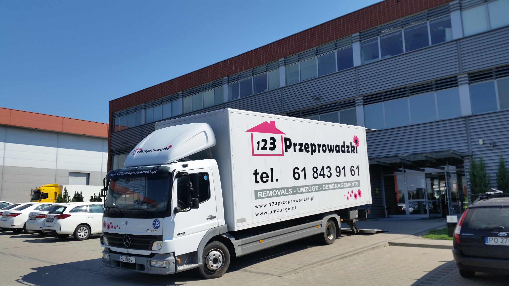 Biała ciężarówka z logo firmy przeprowadzkowej '123 Przeprowadzki' zaparkowana przed nowoczesnym biurowcem w słoneczny dzień, z widocznymi samochodami osobowymi w tle.