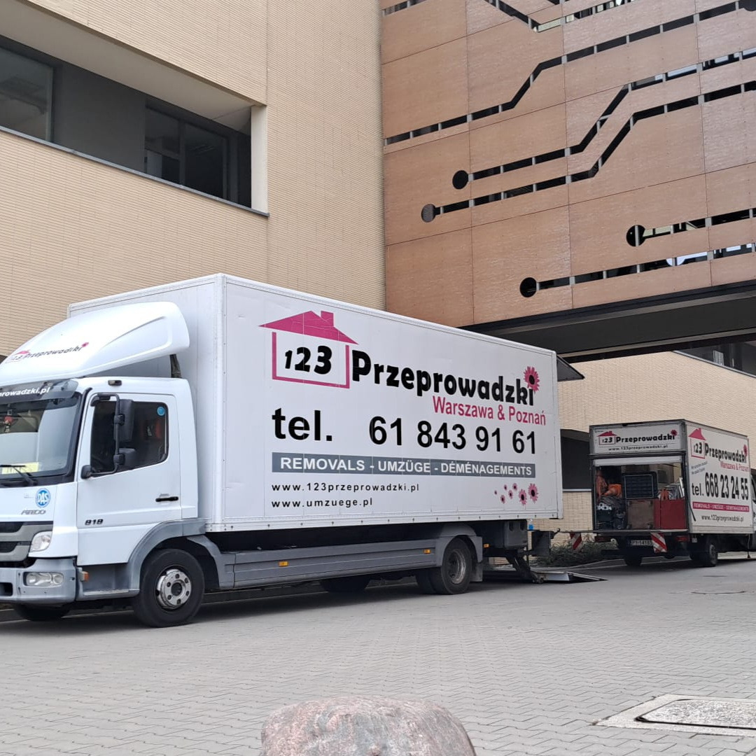 Dwa białe samochody dostawcze firmy przeprowadzkowej z logo i numerem telefonu na tle nowoczesnego budynku biurowego. Widok z boku, lekko z dołu.