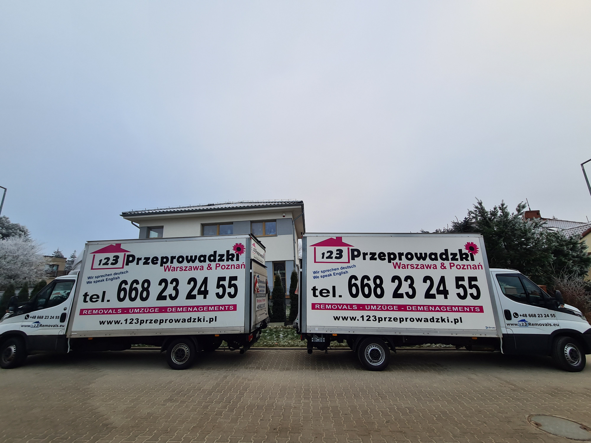 Dwa białe busy z logo firmy przeprowadzkowej '123 Przeprowadzki' na tle budynku. Na busach widoczne numery telefonów i adresy stron internetowych.