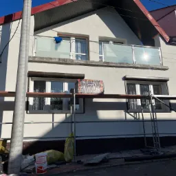 Dom jednorodzinny w trakcie prac elewacyjnych, widoczne rusztowania, materiały izolacyjne i puszki z farbą, na balkonie susząca się niebieska szmatka.