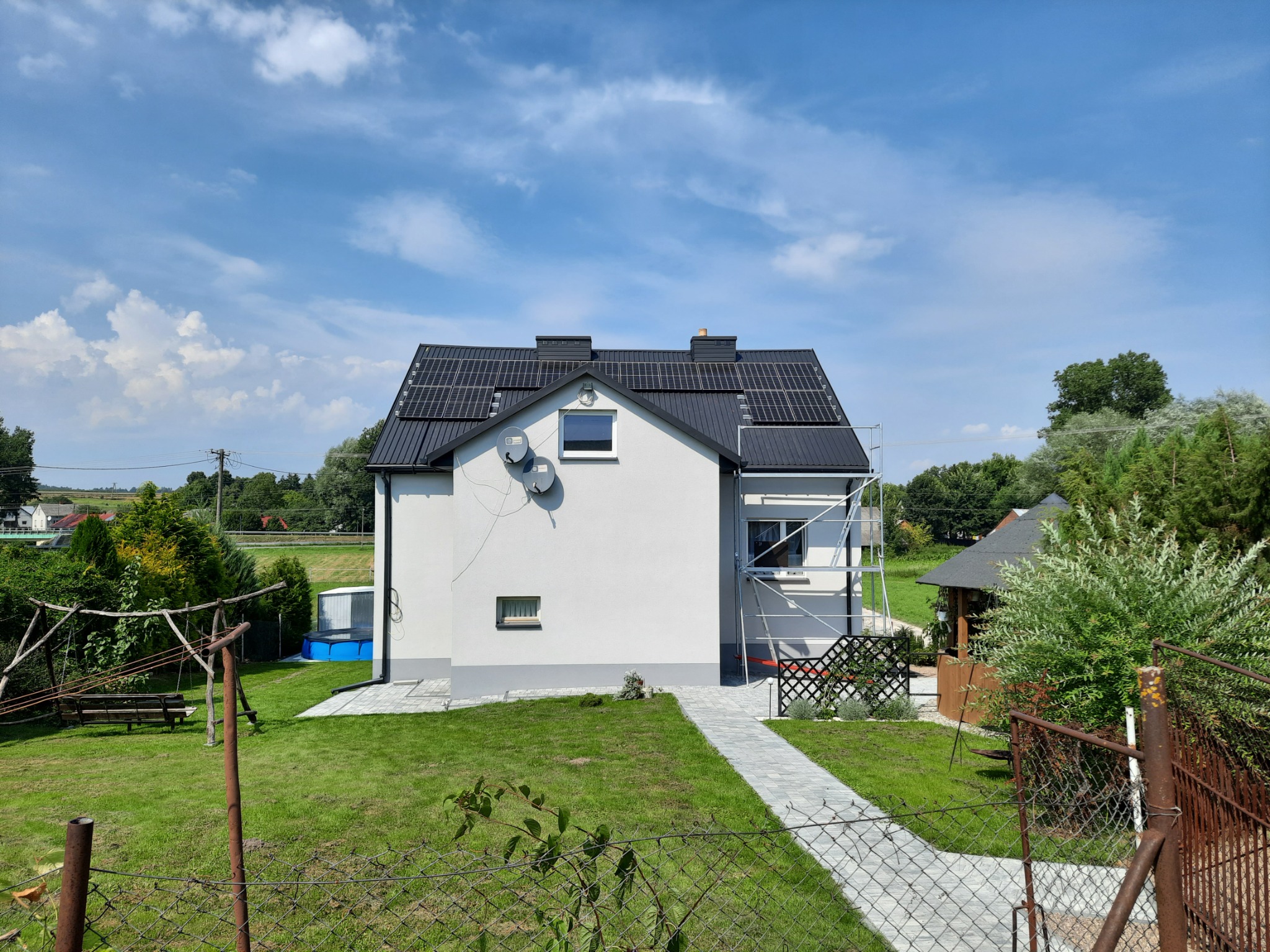 Widok z zewnątrz na dom jednorodzinny z panelami słonecznymi na dachu, częściowo zasłonięty rusztowaniem, na tle błękitnego nieba z chmurami.