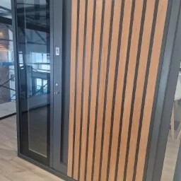 Nowoczesne drzwi wejściowe z przeszklonym panelem i drewnianymi lamelami, szary profil, widoczna klamka i zamek, wnętrze domu w tle.