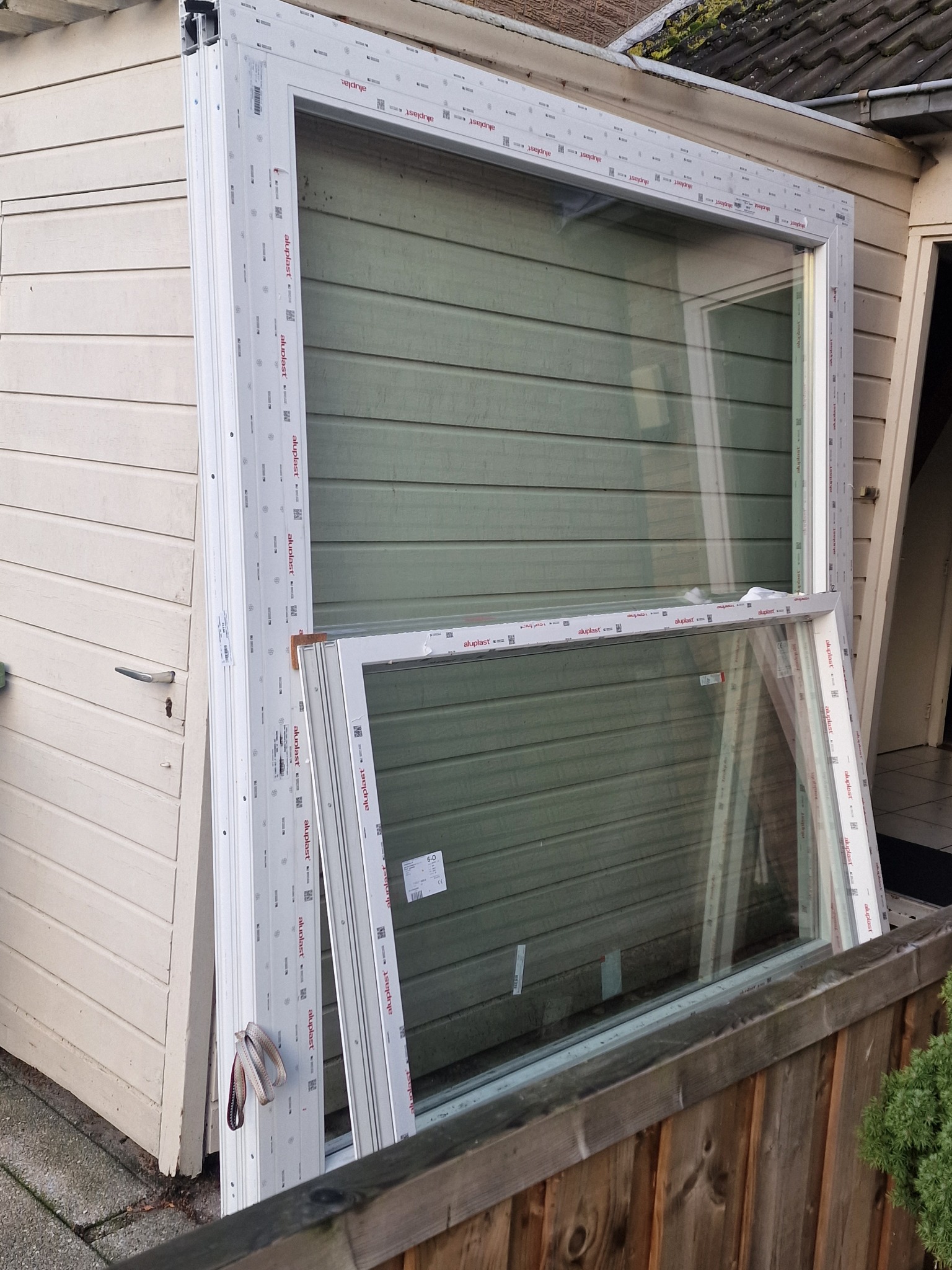 Dwa białe ramy okienne oparte o ścianę budynku, widoczne etykiety producenta na profilach, szare szelki wiszące na jednej z ram.