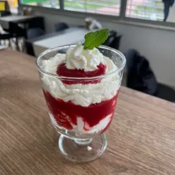 Deser w szklanym pucharku: warstwy bitej śmietany i czerwonego dżemu, udekorowany listkiem mięty na drewnianym blacie stołu w kawiarni.