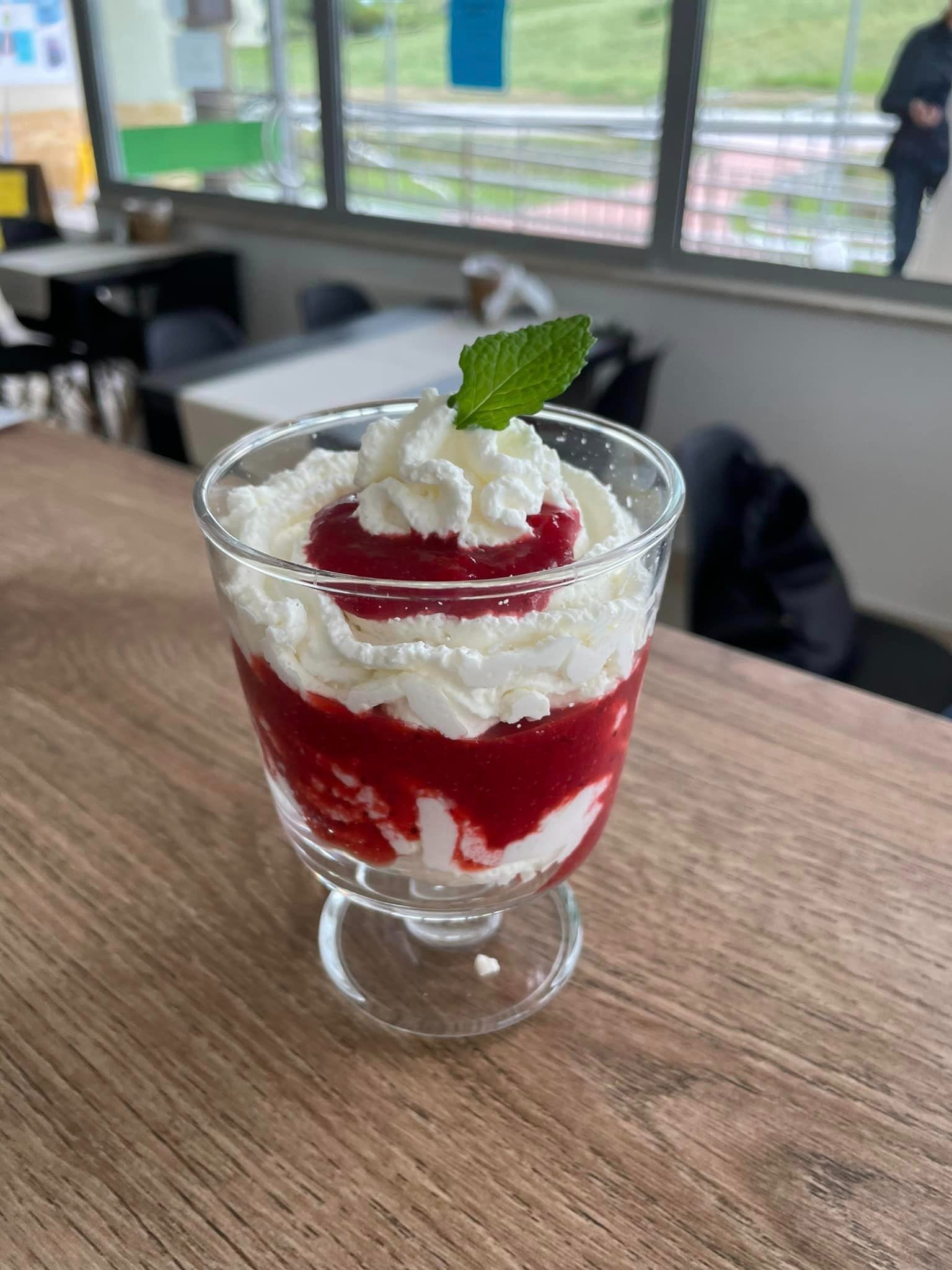 Deser w szklanym pucharku: warstwy bitej śmietany i czerwonego dżemu, udekorowany listkiem mięty na drewnianym blacie stołu w kawiarni.
